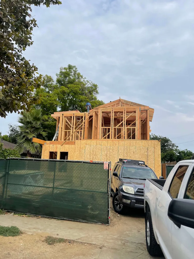 House framing under construction with a green gate and two cars parked nearby.