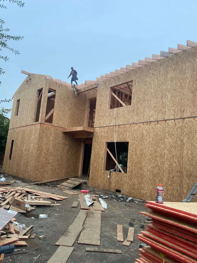 Construction worker on the second story framing a two-story house, with wood materials on the ground.