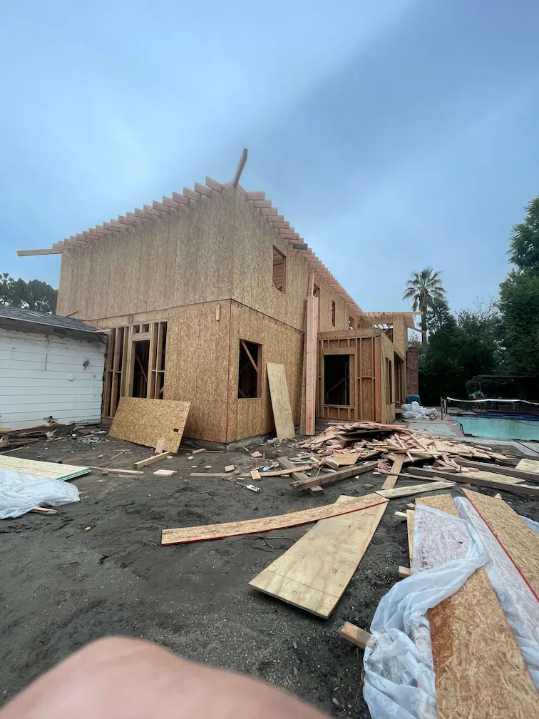Framing in progress for a two-story house with wood on the ground, a pool in the background, and an attachment to an existing property.