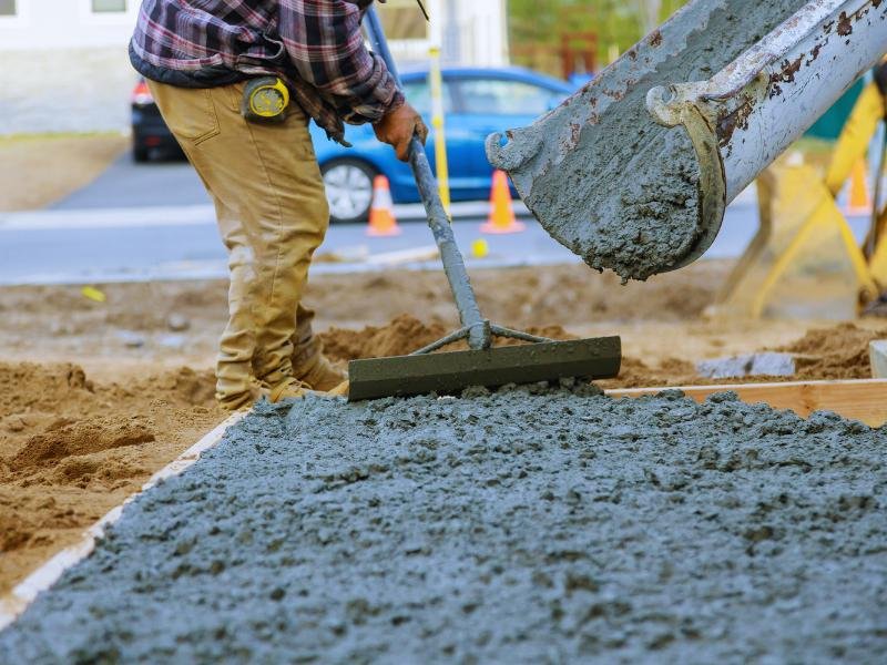 Machine pouring concrete while a construction worker smooths it with a leveling tool.