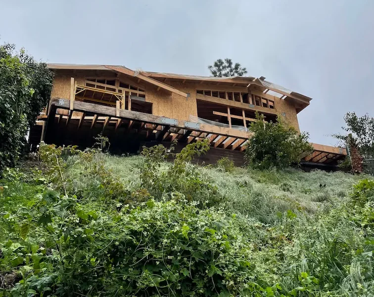 Framing job in progress on a house above a hill, with a greenery-covered slope in the foreground.