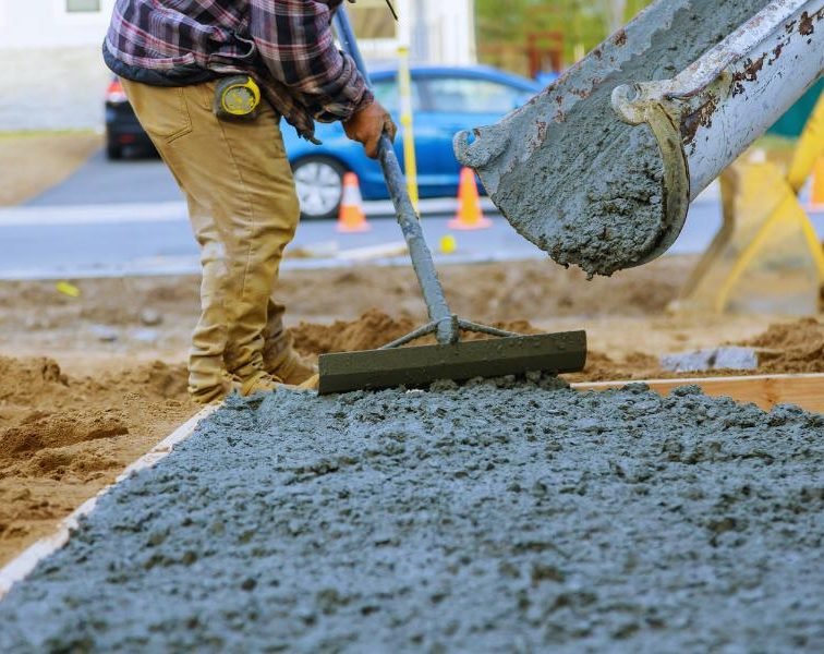 Machine pouring concrete while a construction worker smooths it with a leveling tool.